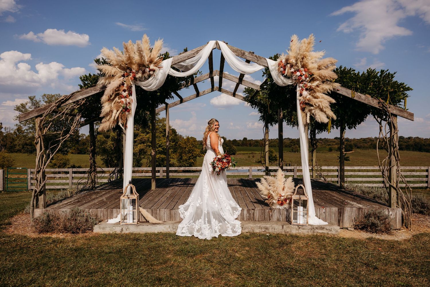 A rustic outdoor wedding arch decorated with pampas grass and white fabric stands in a field at sunset at Carlile Acres.