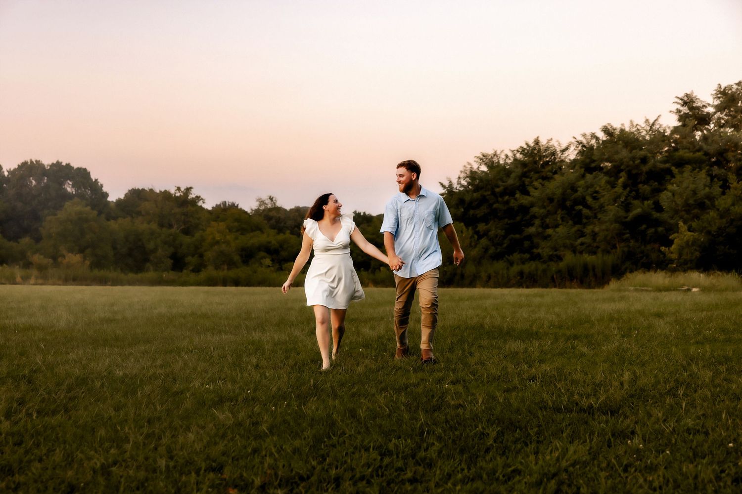 A couple walks hand in hand through a meadow during sunset wearing light summer clothing.
