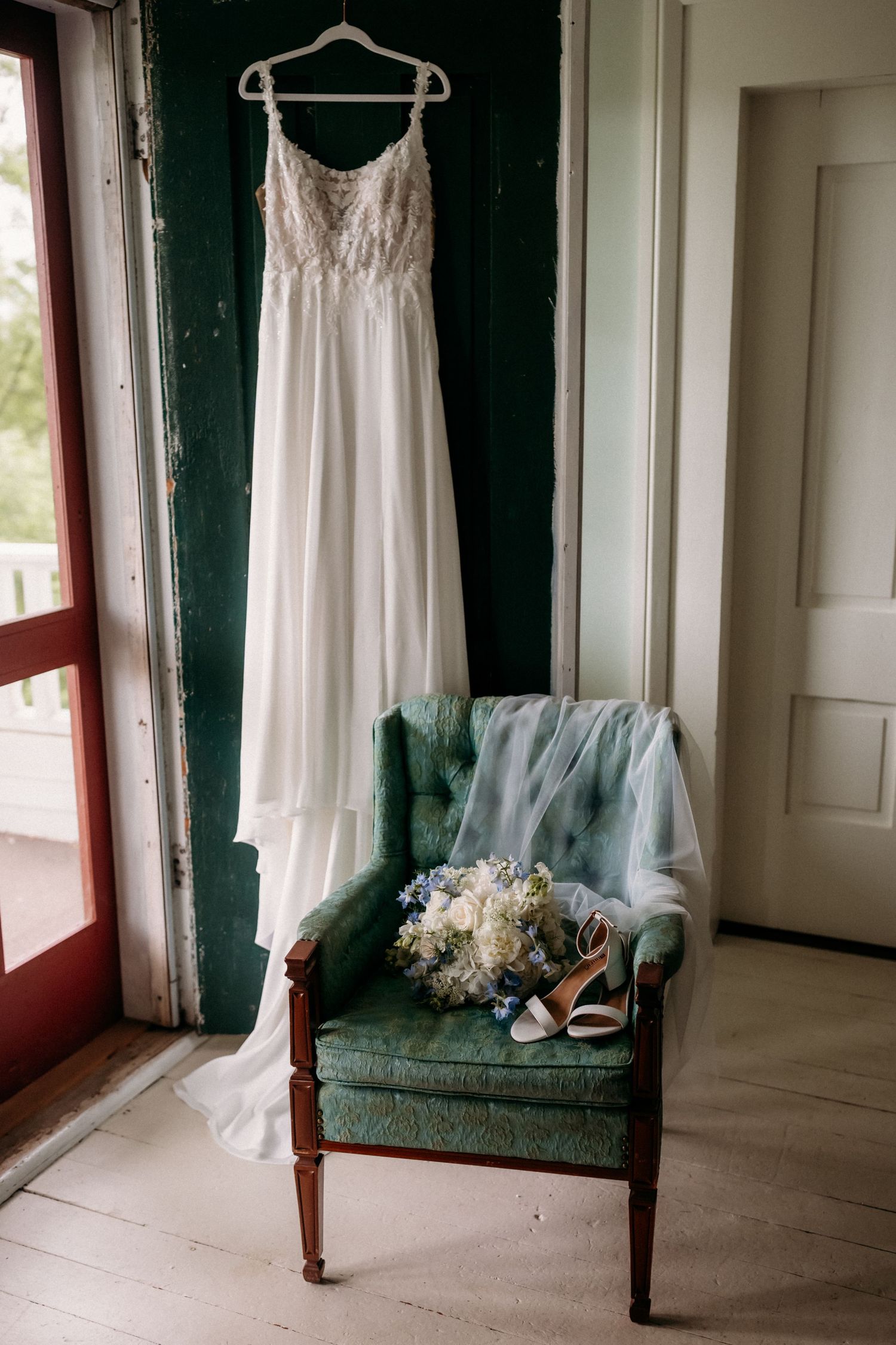 White wedding dress hanging near green chair with bridal bouquet and heels in a vintage room.