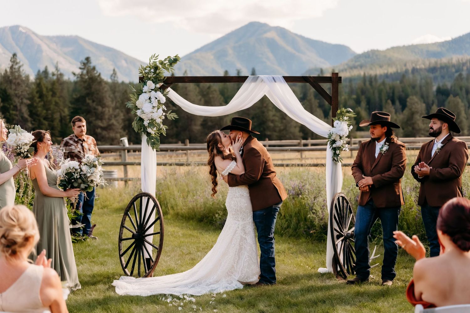 Bride and groom share first kiss at western-themed outdoor altar with wagon wheels and mountain views.
