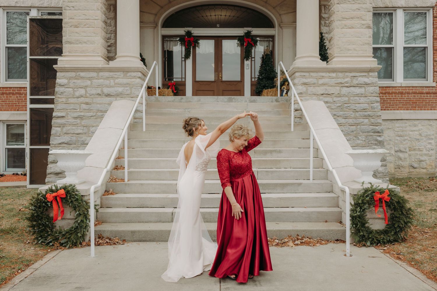 Bride in white gown twirls with woman in red dress outside stone building decorated with Christmas wreaths.