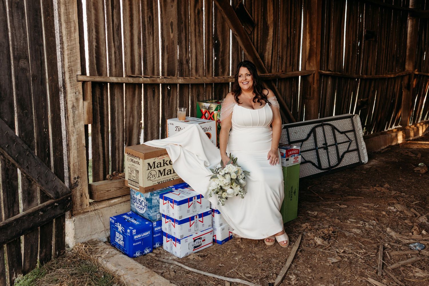 Bride in white gown poses with beer cases and bouquet inside a rustic barn at wedding venue.