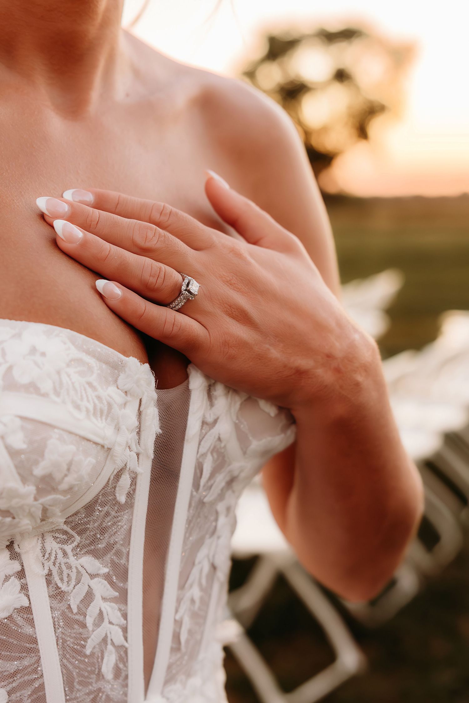Bride in lace strapless gown wearing a diamond engagement ring at golden hour sunset.