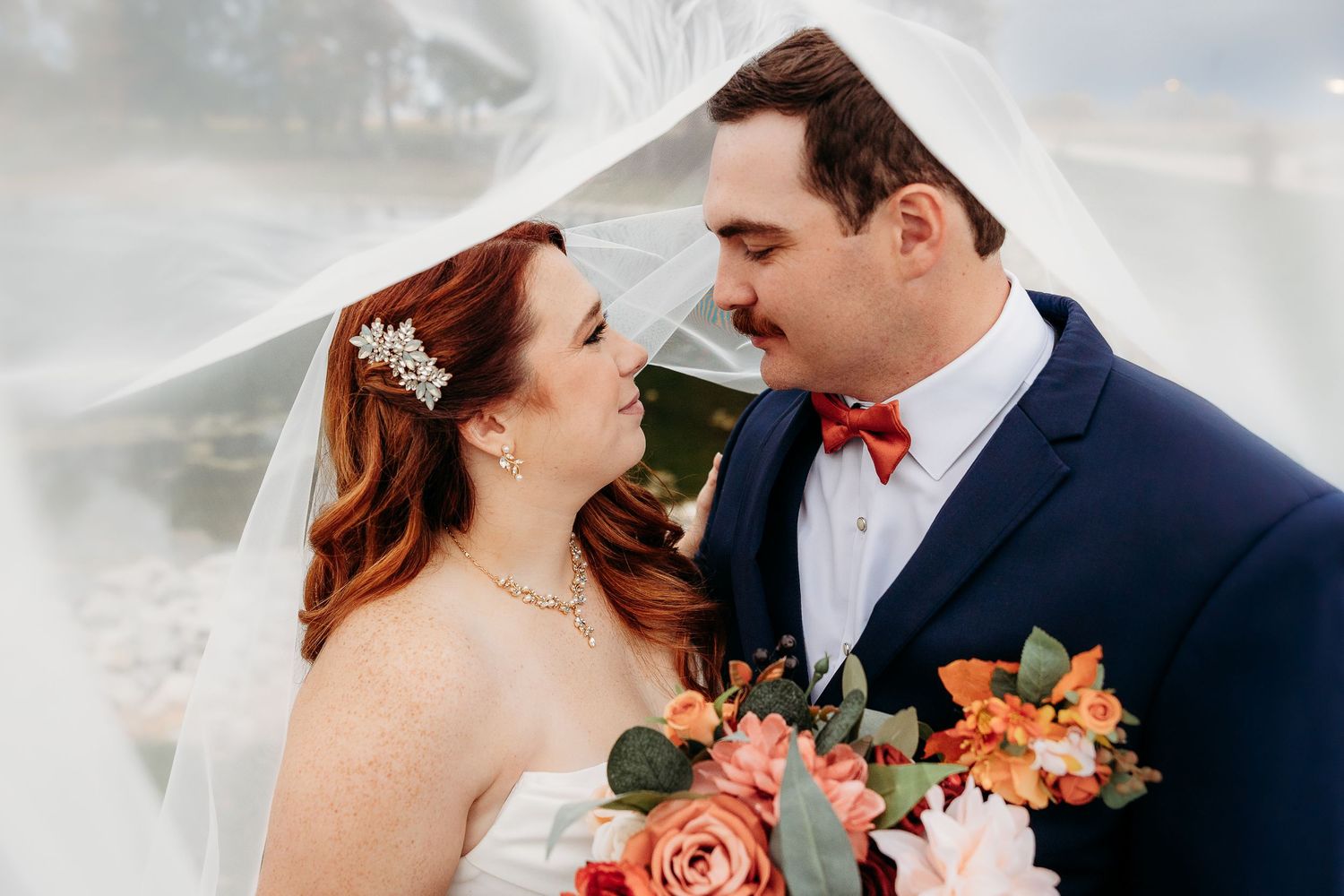 Bride with red hair and groom in navy suit share a tender moment under flowing veil with colorful bouquet.