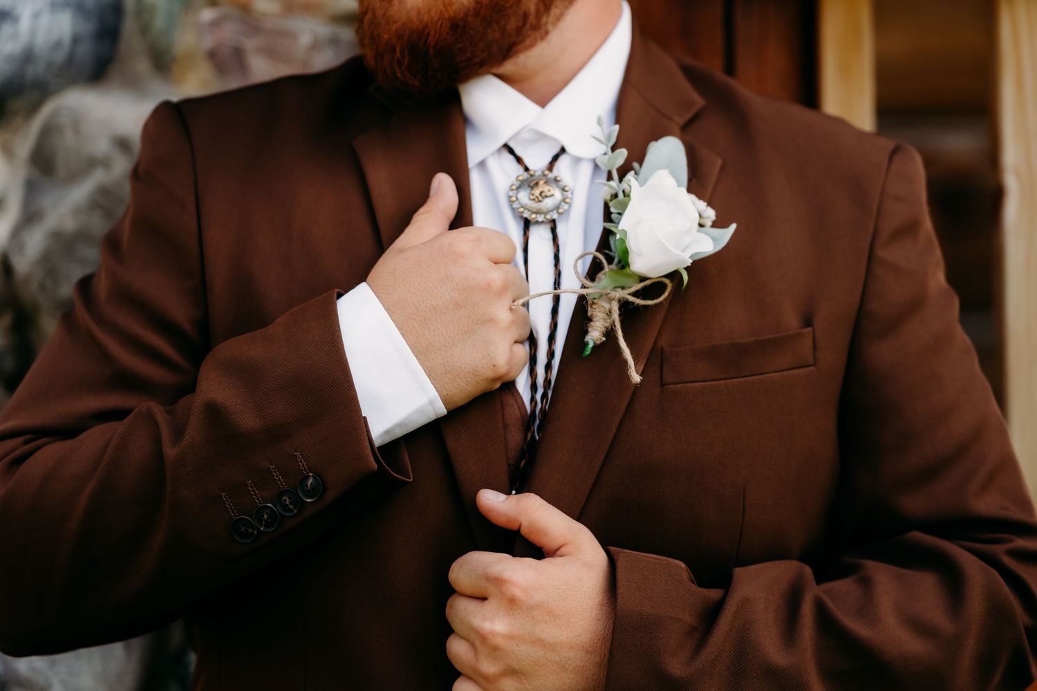 Groom in brown suit adjusts bolo tie with white rose boutonniere at rustic western-themed wedding ceremony.
