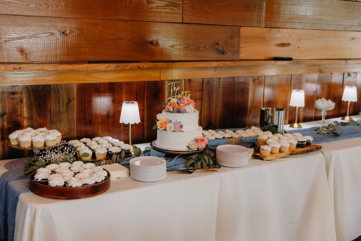 Elegant wedding dessert table with tiered cake, cupcakes, and small lamps against rustic wood wall.