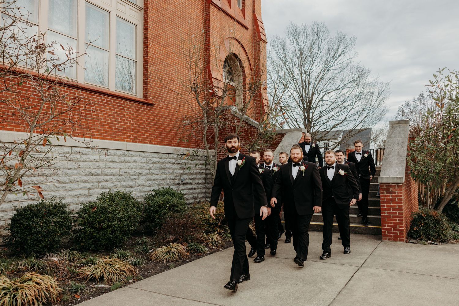 Groom leads groomsmen in black tuxedos walking along brick church exterior pathway on wedding day.