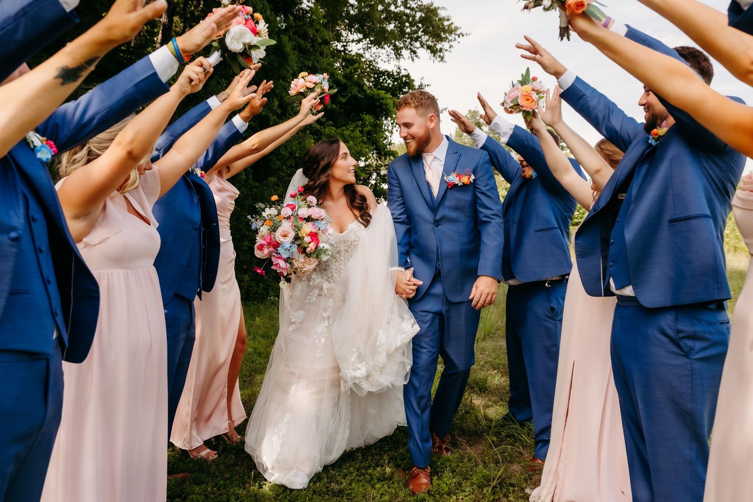 Bride and groom walk through a tunnel of raised hands formed by wedding party in blue suits and blush gowns outdoors.