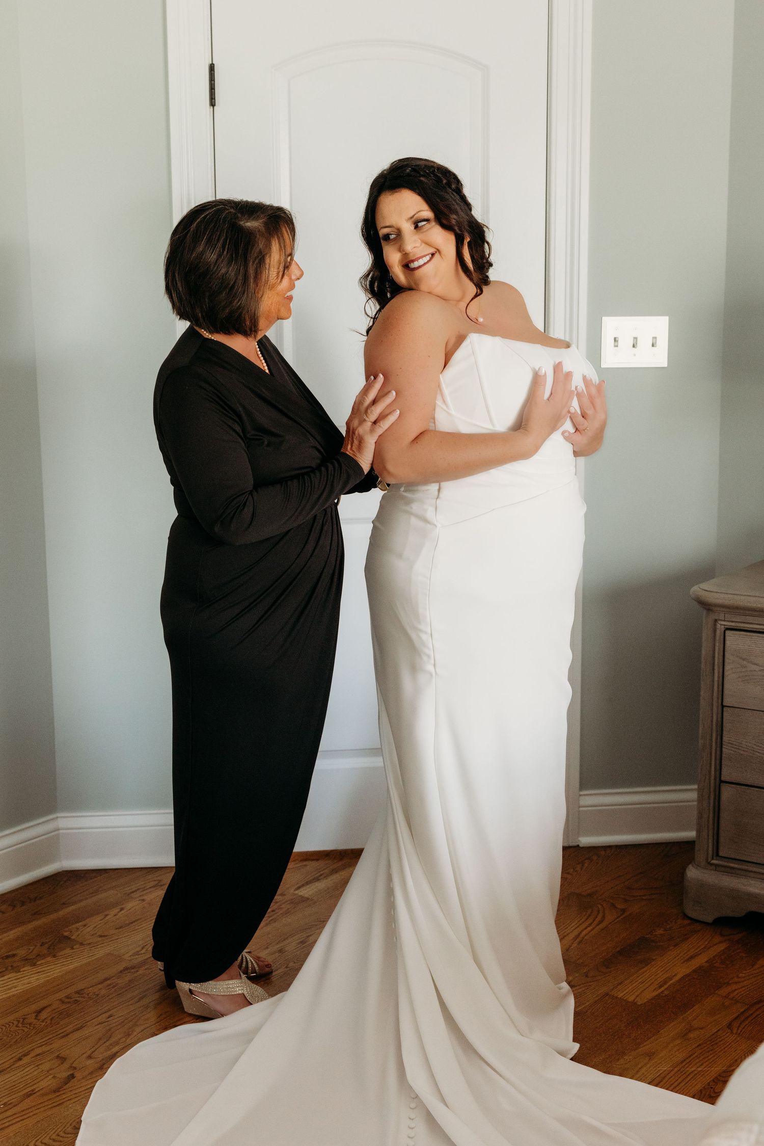 Mother helping bride into fitted white wedding gown in a softly lit dressing room.