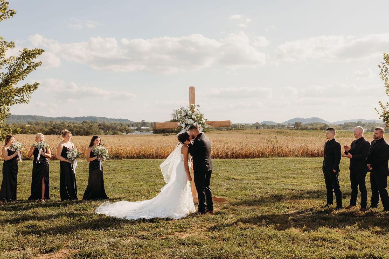 Bride and groom kiss at outdoor wedding ceremony with bridesmaids in black dresses and groomsmen against scenic field backdrop.