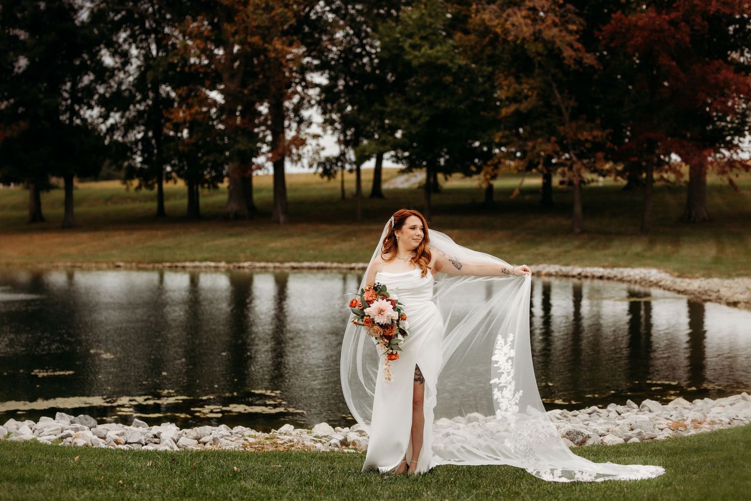 Dramatic bridal portraits by a lake with flowing veil and fall foliage in background.