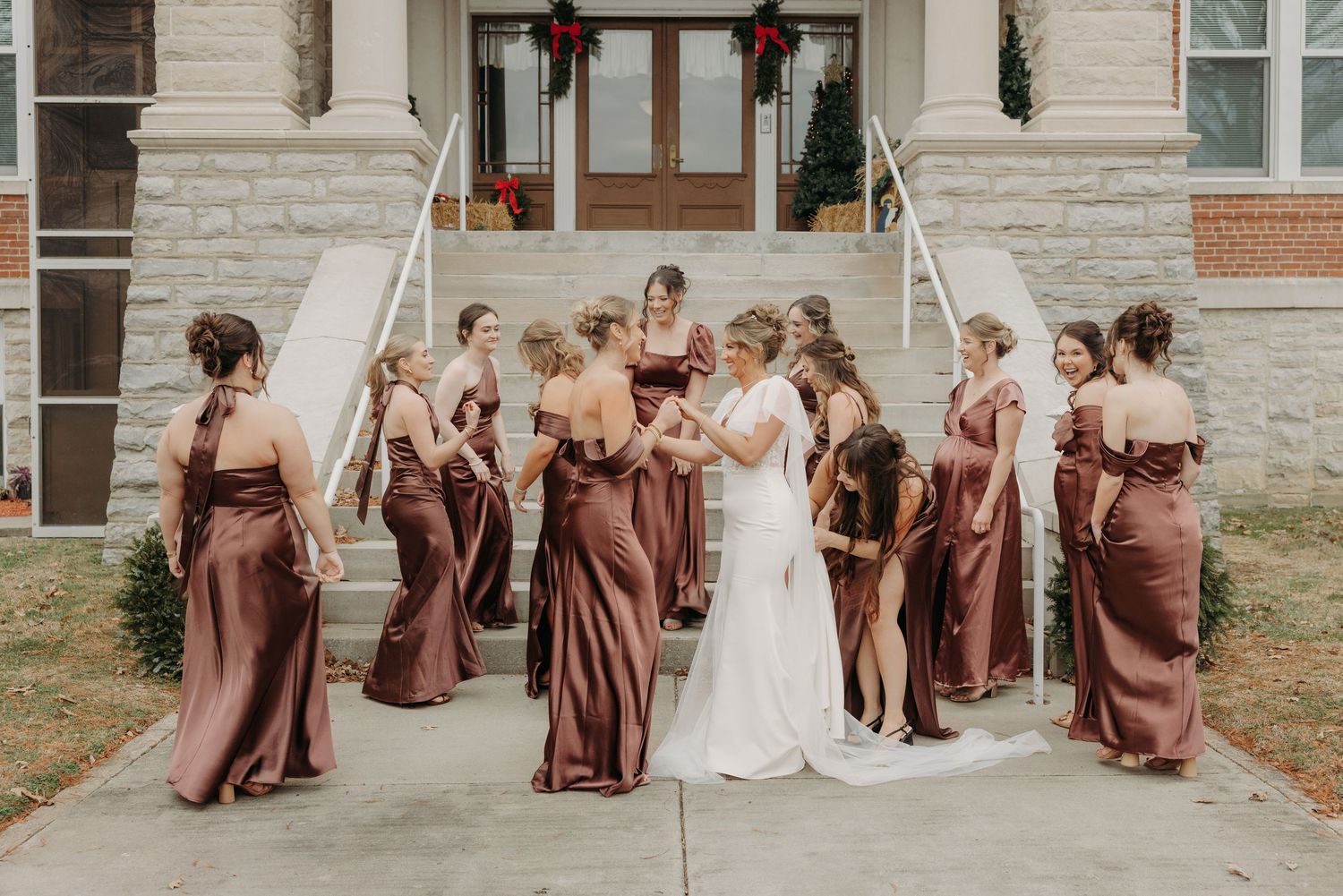 Bride surrounded by bridesmaids in elegant rust-colored satin gowns on stone steps of a historic building.
