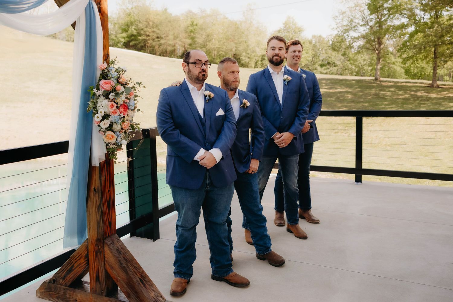 Groom and groomsmen in navy blue suits and brown boots standing at outdoor wedding ceremony altar.
