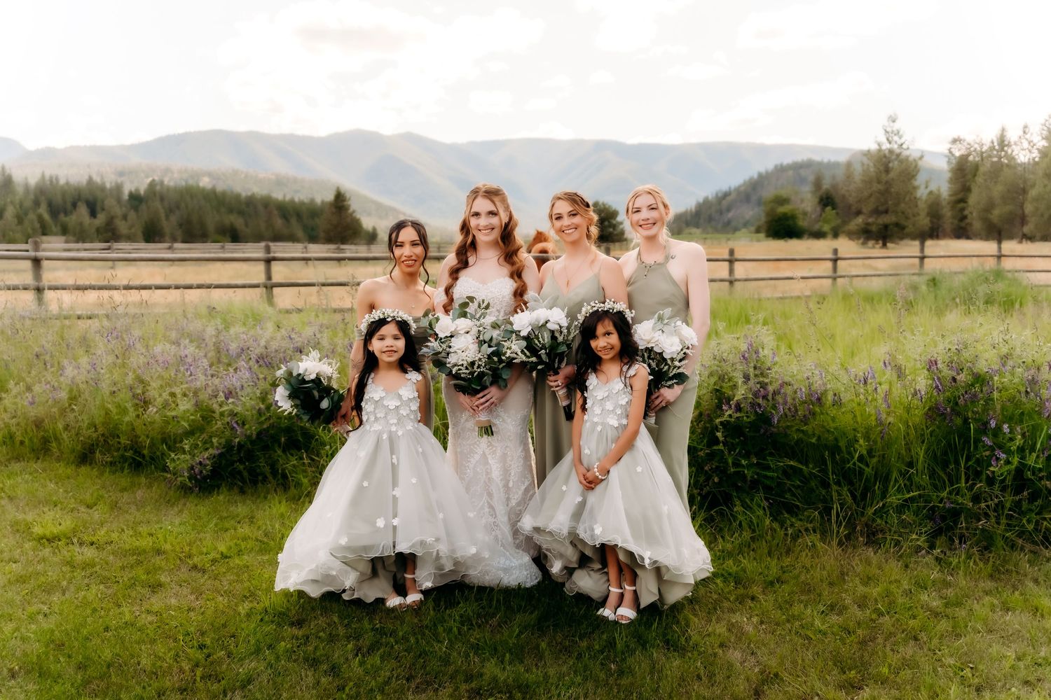 Bride with bridesmaids and flower girls holding white bouquets in a mountain meadow setting.