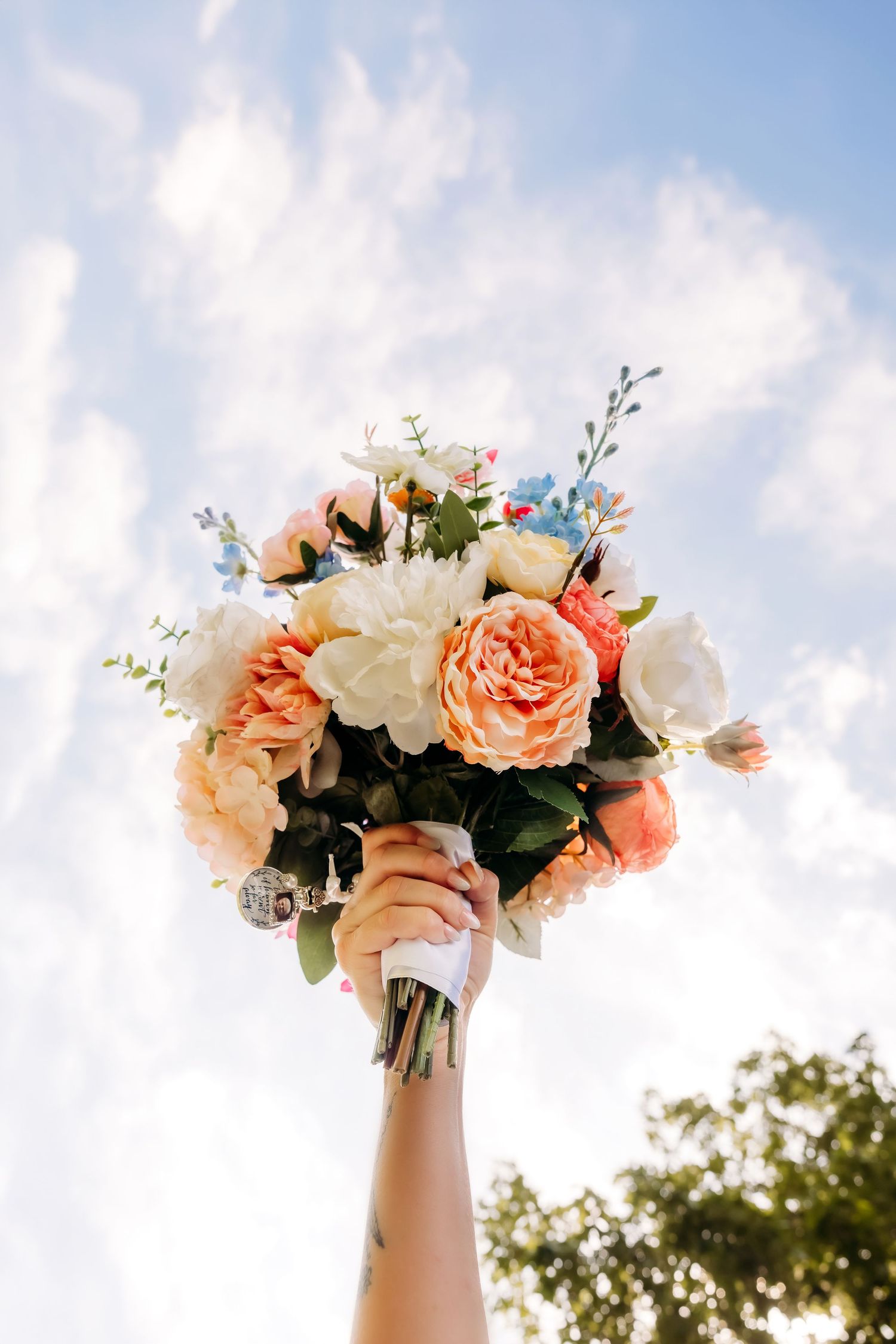 Hand holding a vibrant bridal bouquet of peach, white, and blue flowers raised against a bright cloudy sky.