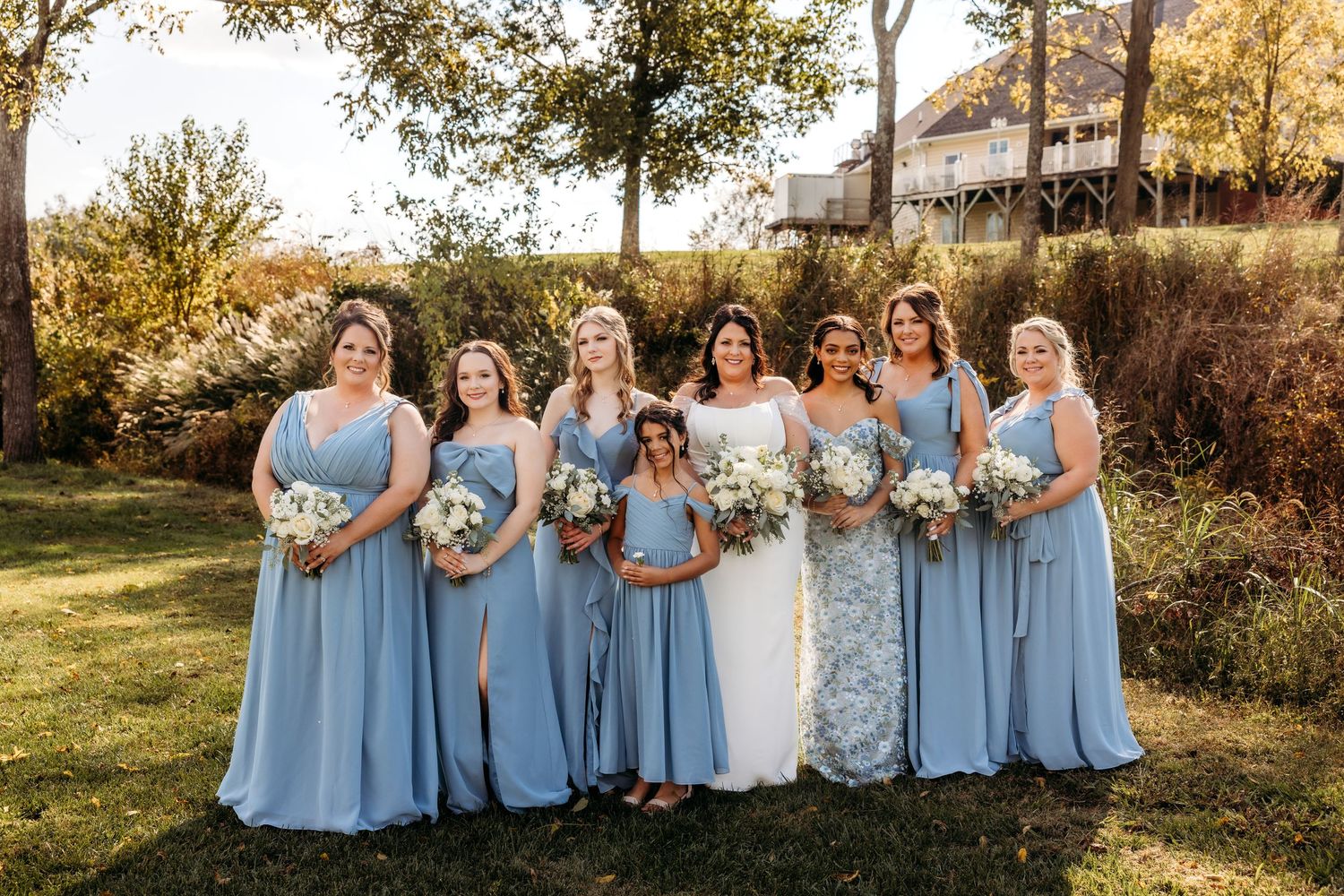 Bride with bridesmaids in dusty blue gowns holding white bouquets outdoors on a sunny autumn day.