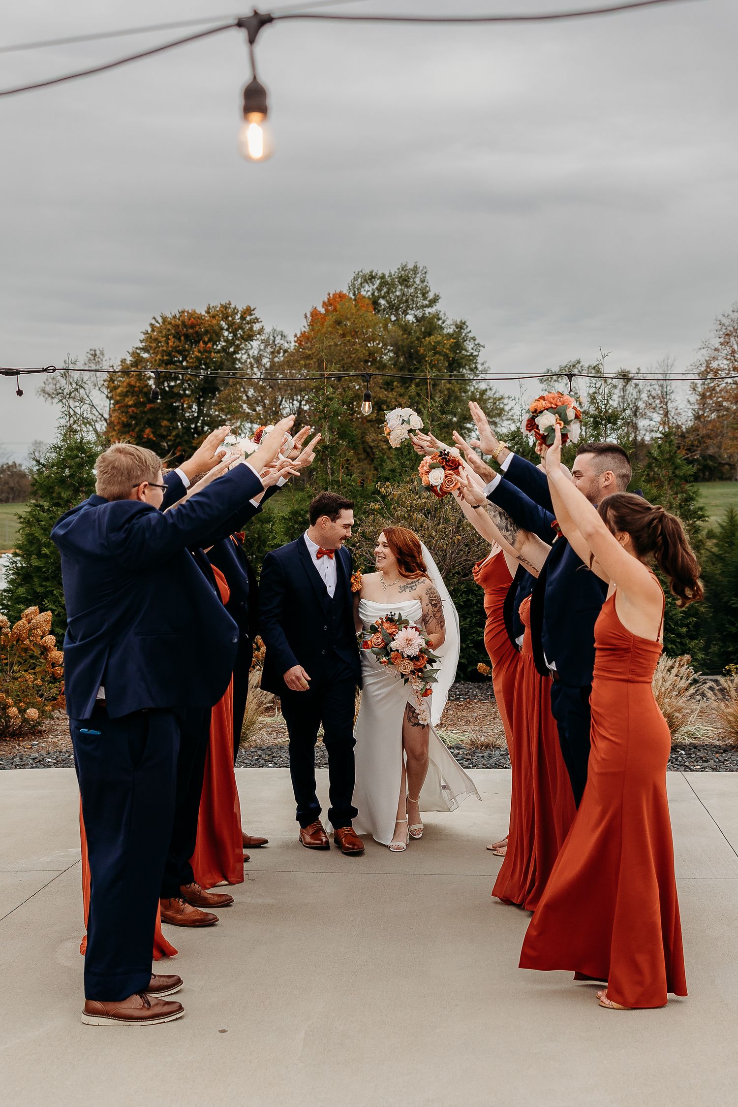 Wedding party raises bouquets forming arch as bride and groom walk through outdoor celebration exit.