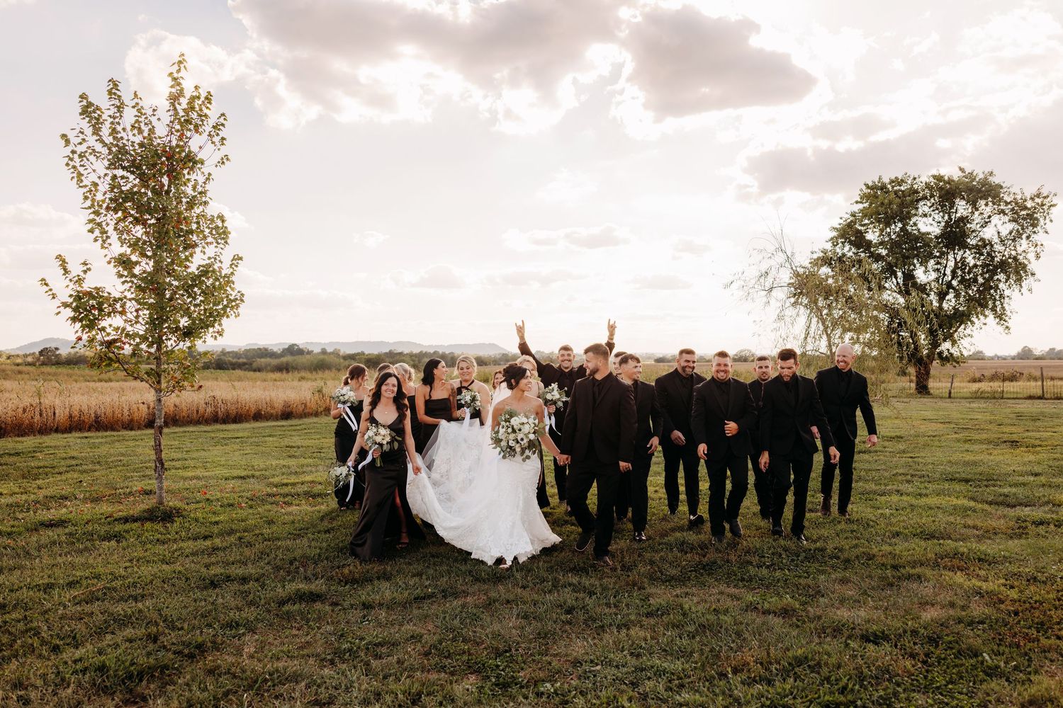 Wedding party in black attire celebrates with bride and groom on a green field at golden hour.