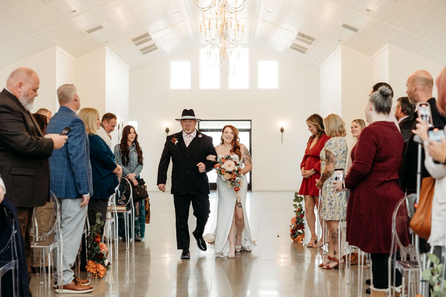 Bride walks down white chapel aisle escorted by father in black suit before wedding ceremony guests.