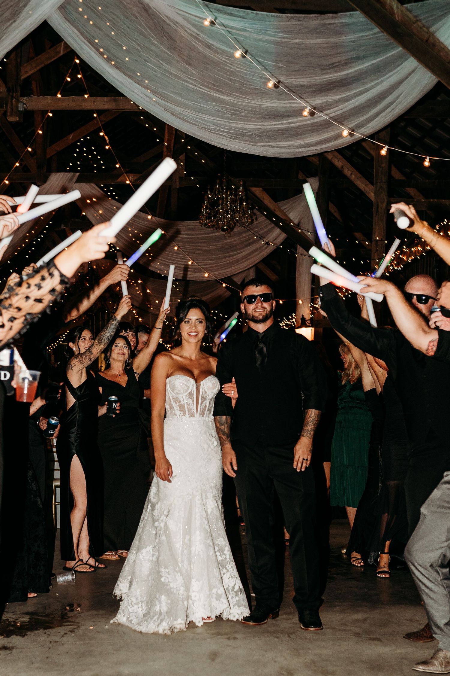 Newlyweds exit wedding reception through a tunnel of glowing light sticks held by cheering guests in a barn.