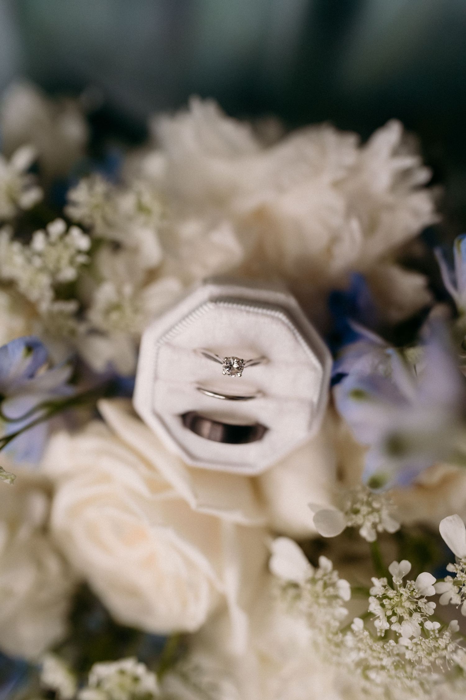 Diamond engagement ring and wedding band displayed in white velvet box surrounded by blue and white floral bouquet.
