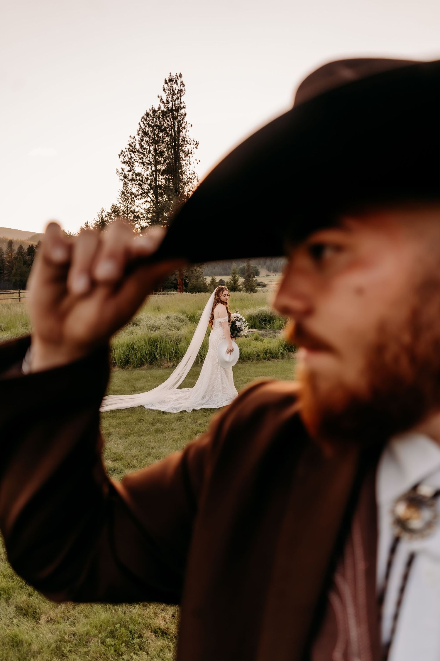 Groom in black cowboy hat and brown jacket tips his hat while bride in white gown stands in background.
