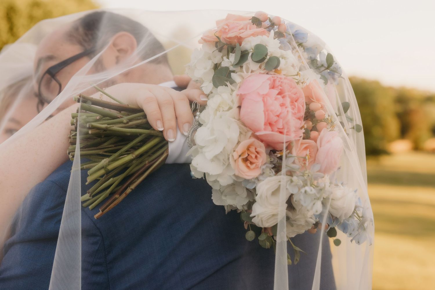 Bride and groom sharing intimate moment under veil, holding pink and white wedding bouquet.