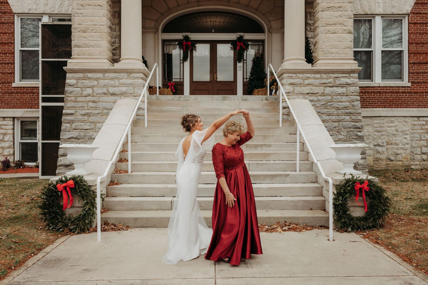 Mother of the bride dances in burgundy dress celebrating on stone steps with bride.