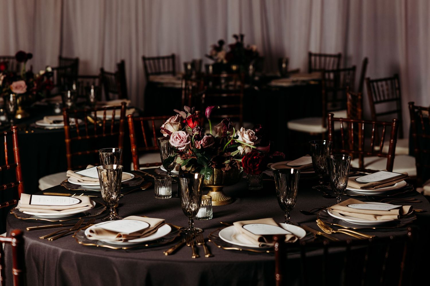 Elegant dark wedding reception tables with gold cutlery, white plates, and lush floral centerpieces.