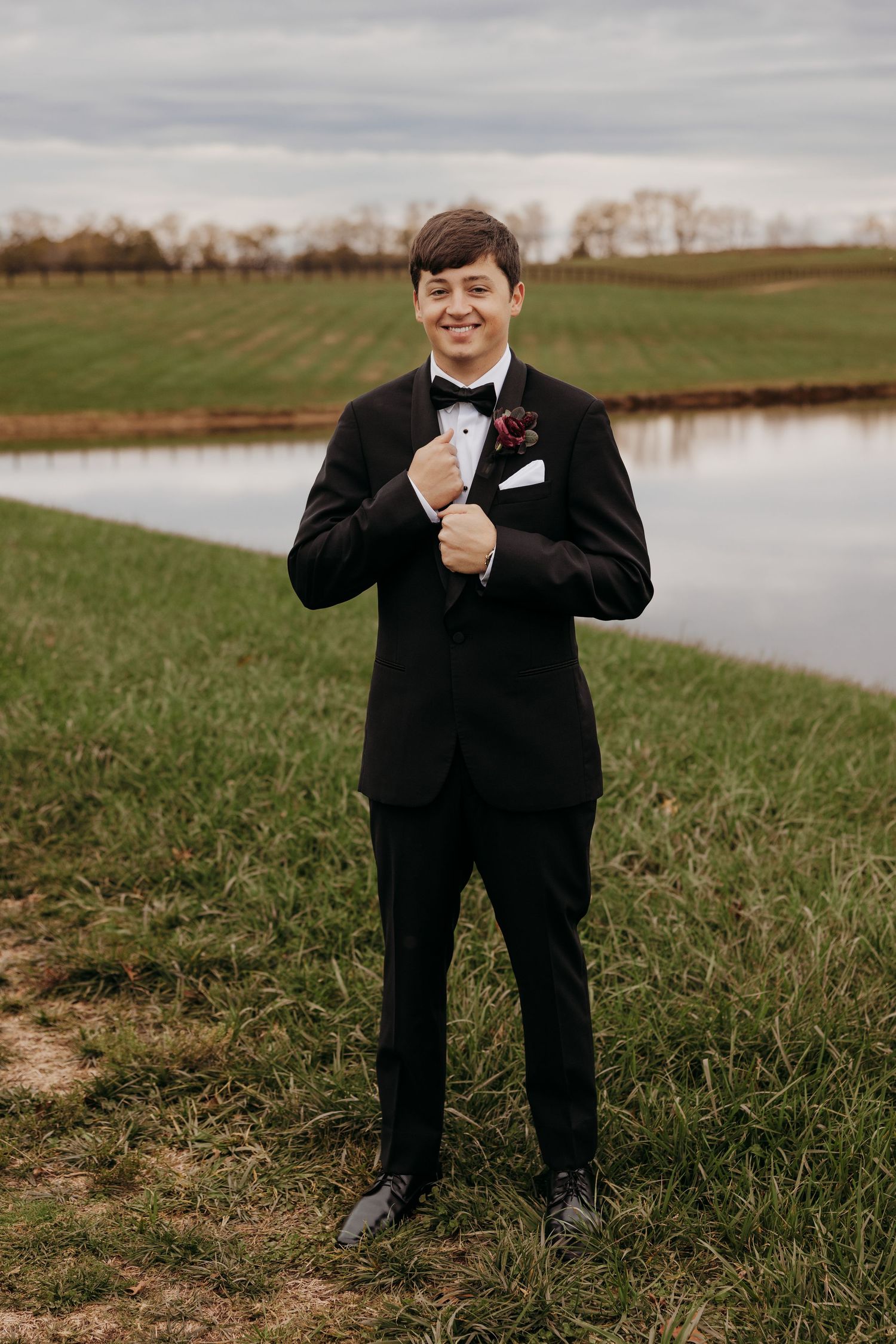 Smiling groom in black tuxedo with bow tie and boutonniere standing by a pond in a green field.