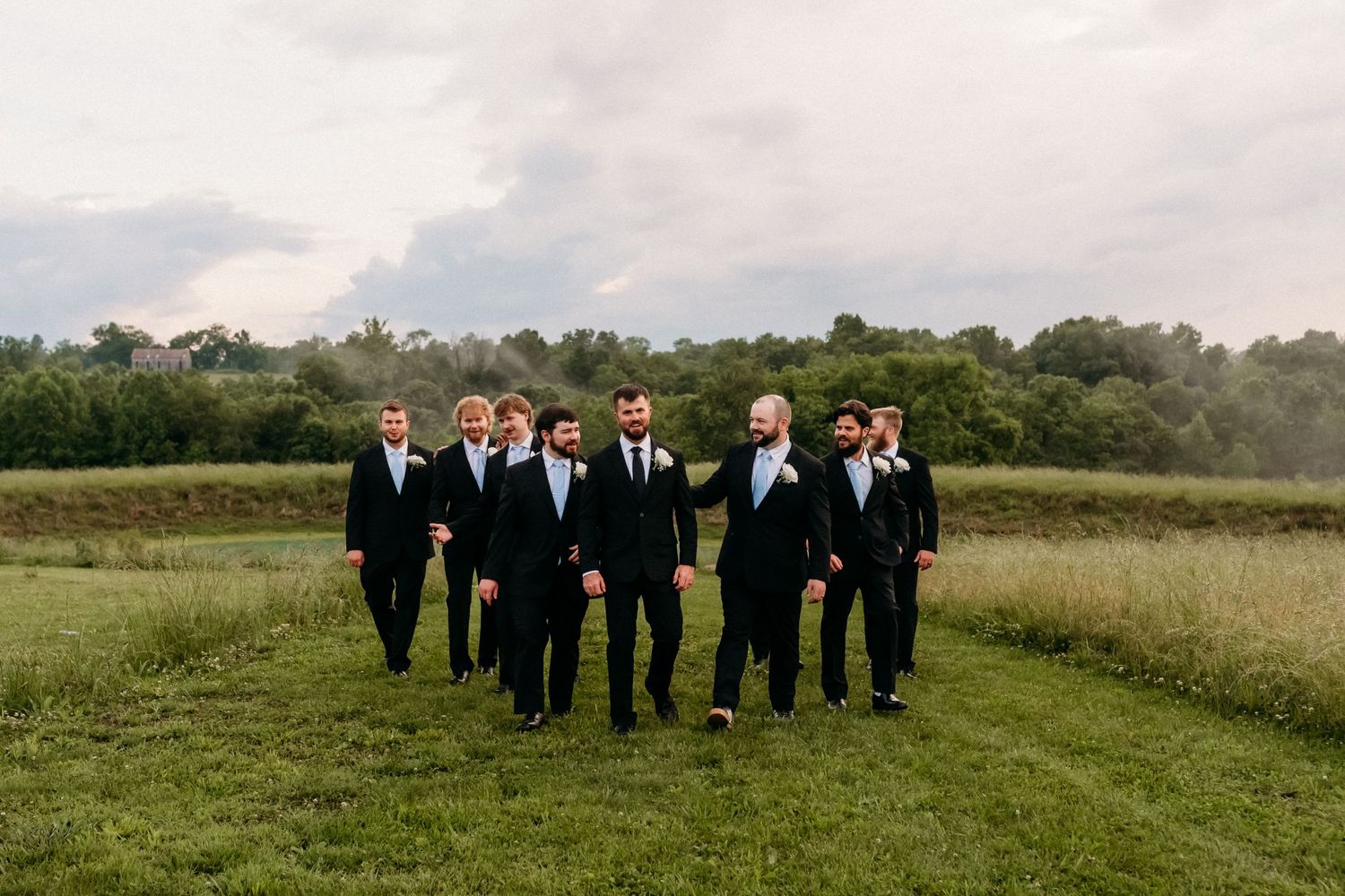 Groomsmen in black tuxedos walking together across a green field at an outdoor wedding celebration.