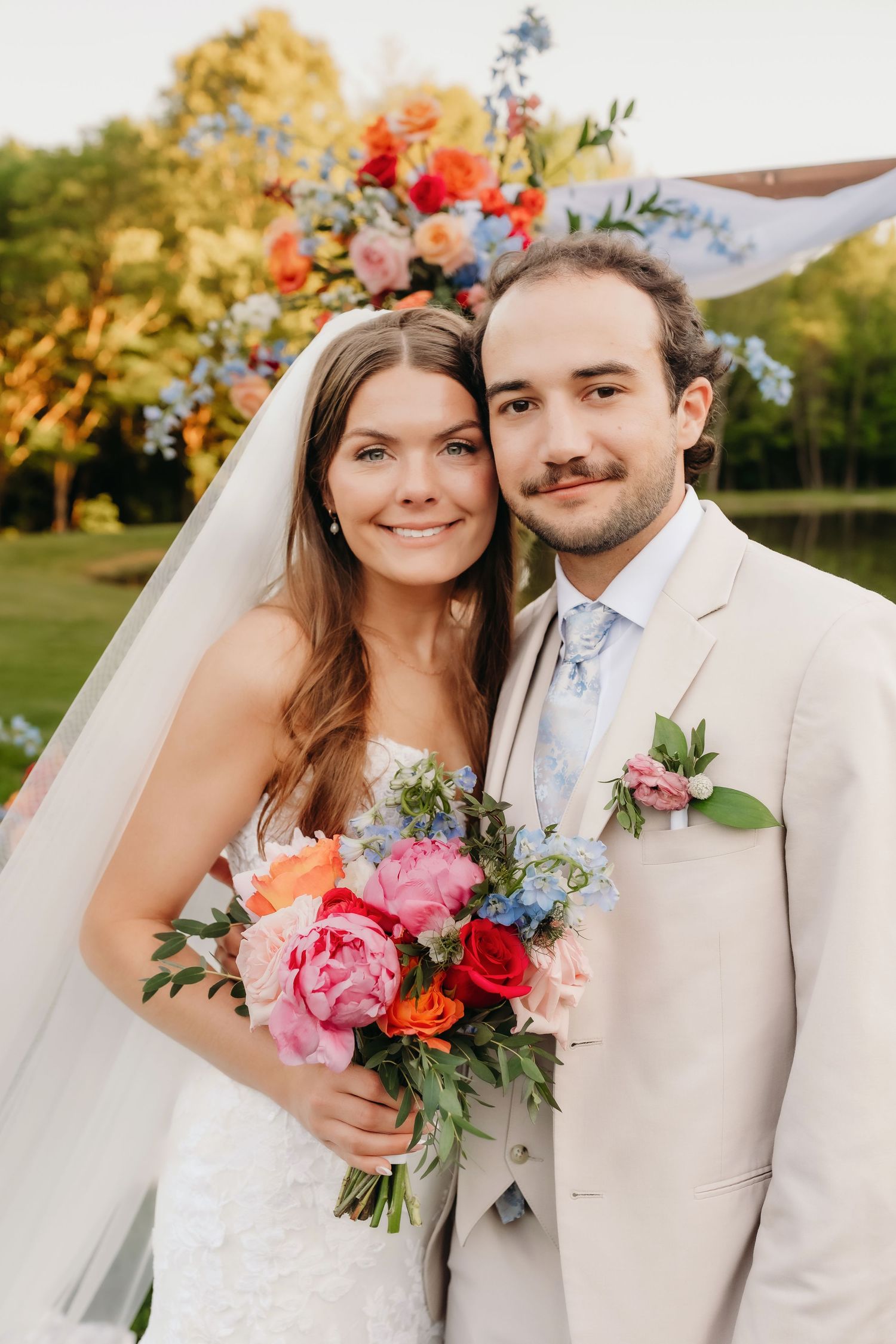 Bride and groom smiling together holding colorful bouquet with floral arch backdrop at outdoor wedding ceremony.