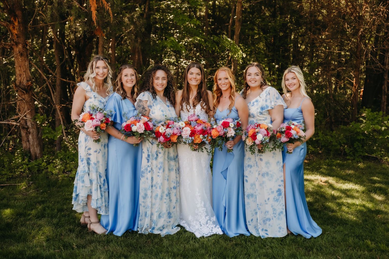 Bride and bridesmaids in blue dresses holding colorful bouquets pose outdoors in front of lush green trees.