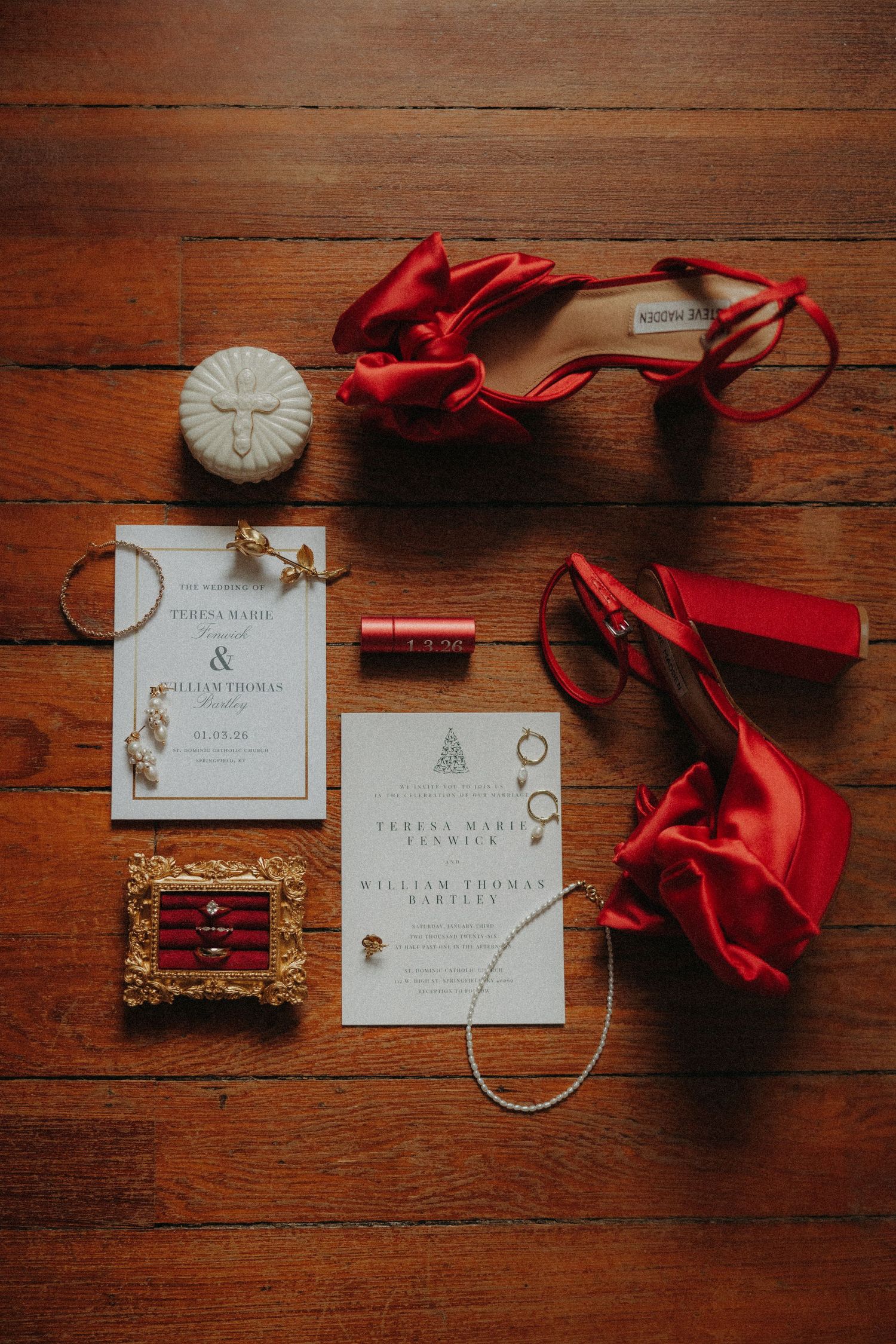 Elegant wedding flat lay with red heels, gold ring box, pearl necklace, lipstick, and invitation suite on wood floor.