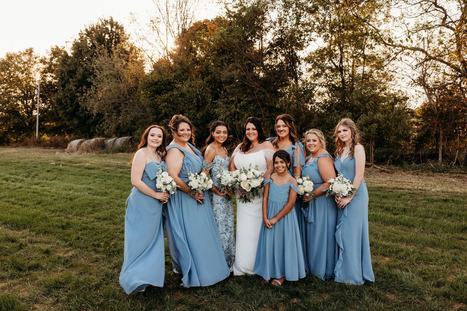 Bride with six bridesmaids in dusty blue gowns holding white bouquets, posing in a sunlit field at golden hour.