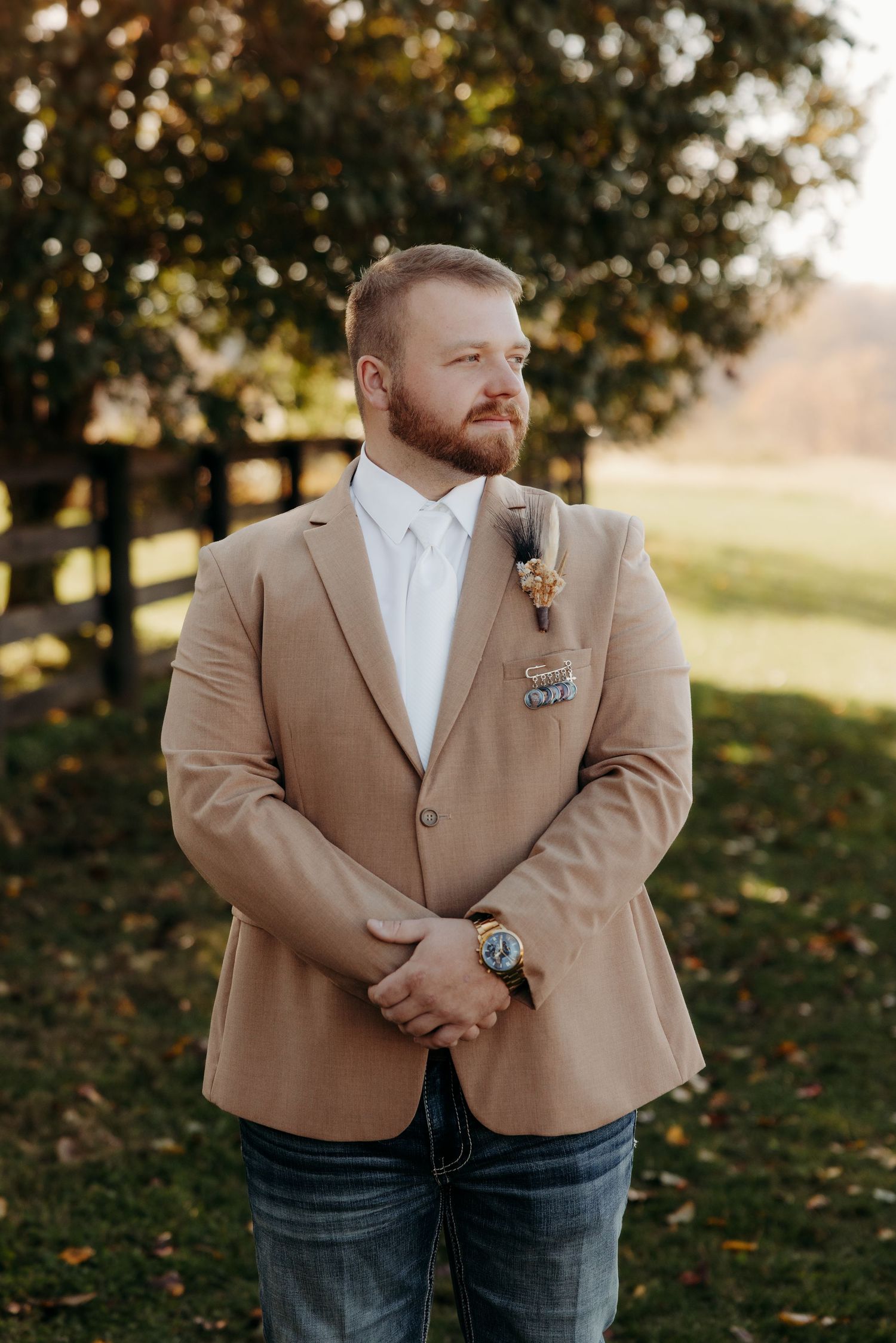 Groom in tan blazer with boutonniere and jeans standing outdoors at fall wedding venue.