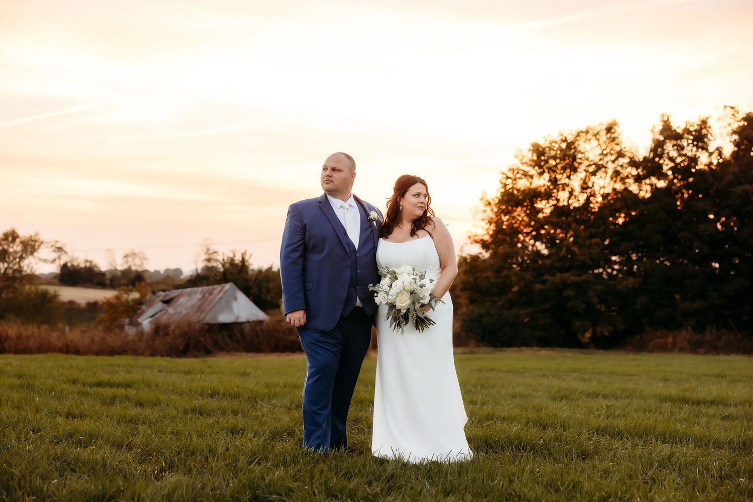 Bride and groom stand in golden sunset field, she holds white bouquet, rustic barn in background.