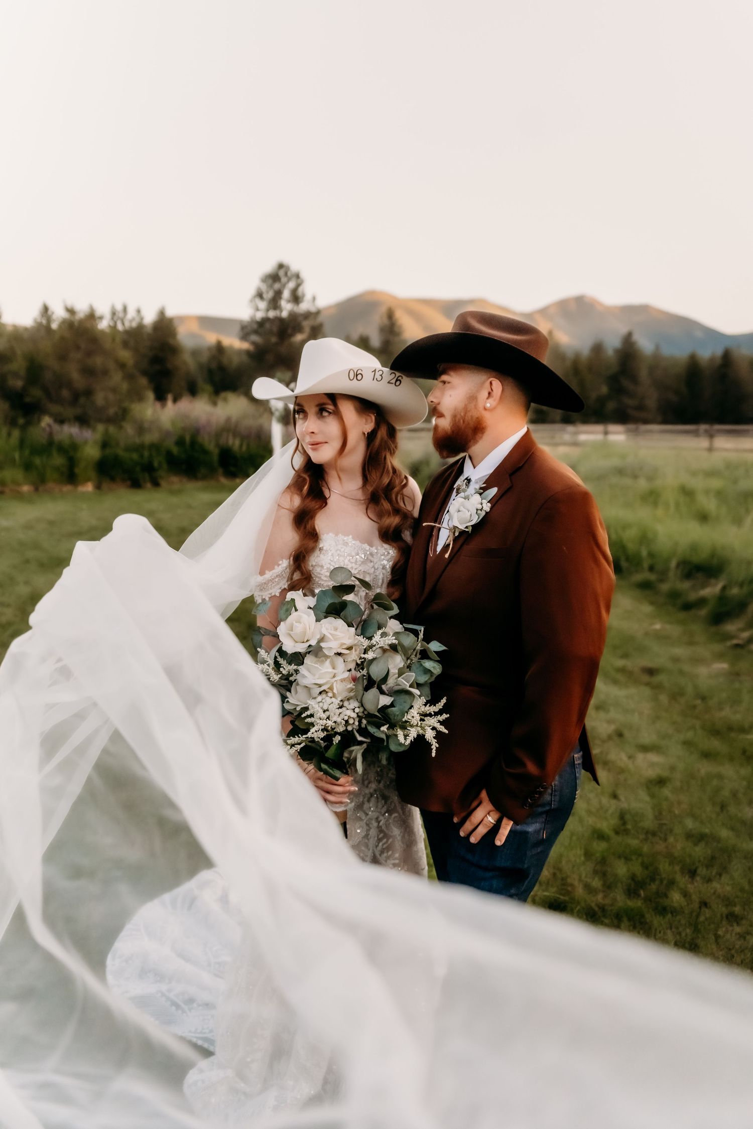 Bride and groom in cowboy hats pose outdoors with veil flowing in wind, mountain backdrop at rustic western wedding.