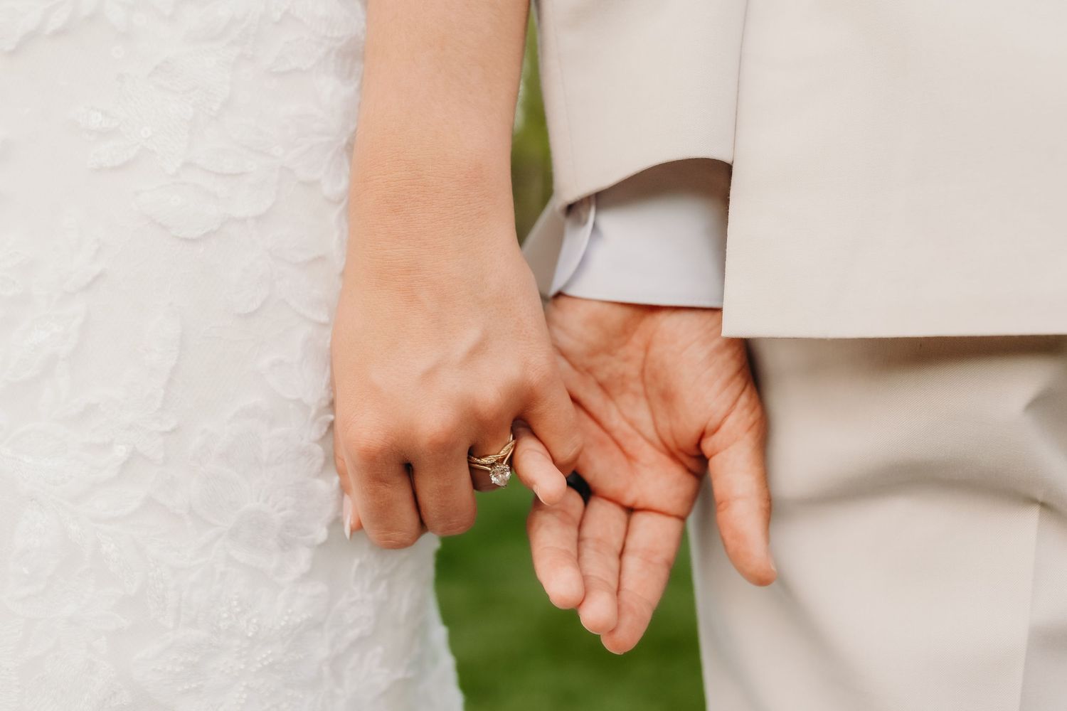 Close-up of bride and groom holding hands showing wedding rings in white lace and beige suit.