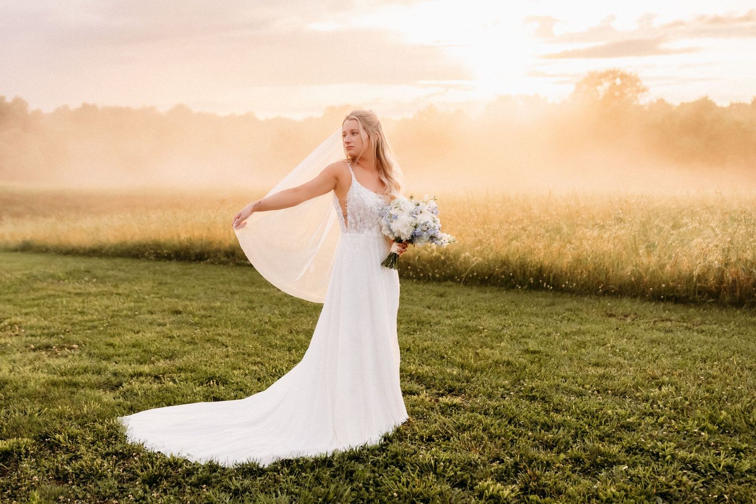 Bride in white gown holding bouquet at golden sunset in misty field.