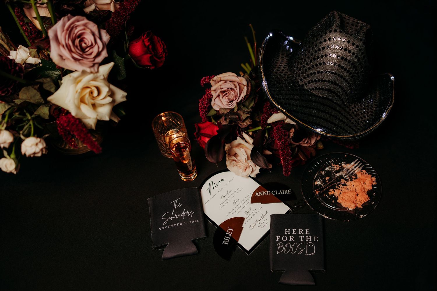 Dark moody wedding flat lay with red roses, champagne flutes, menus, and decorative fan on black background.