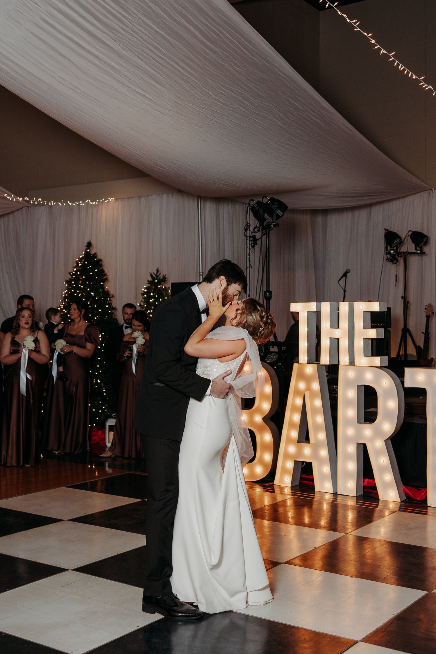 Bride and groom share first dance kiss in front of marquee letter sign on black and white checkered dance floor.