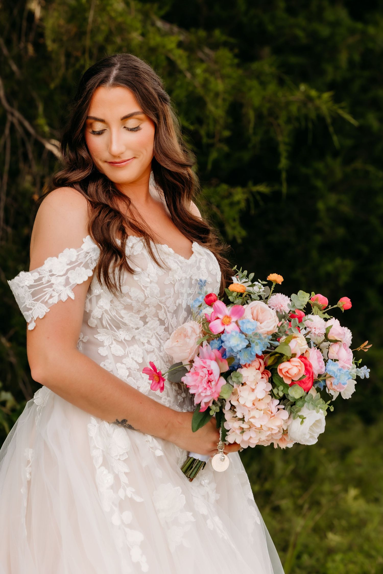 Bride in off-shoulder lace wedding gown holds colorful pink and blue floral bouquet outdoors.