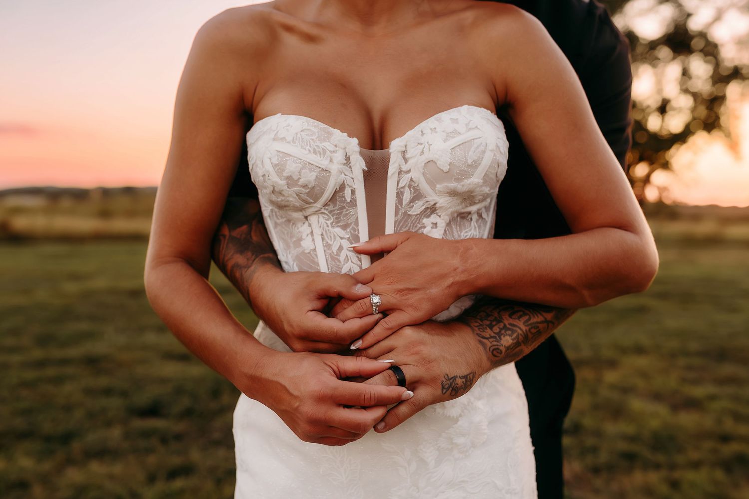 Bride in lace corset wedding dress embraced by groom at sunset on green field.