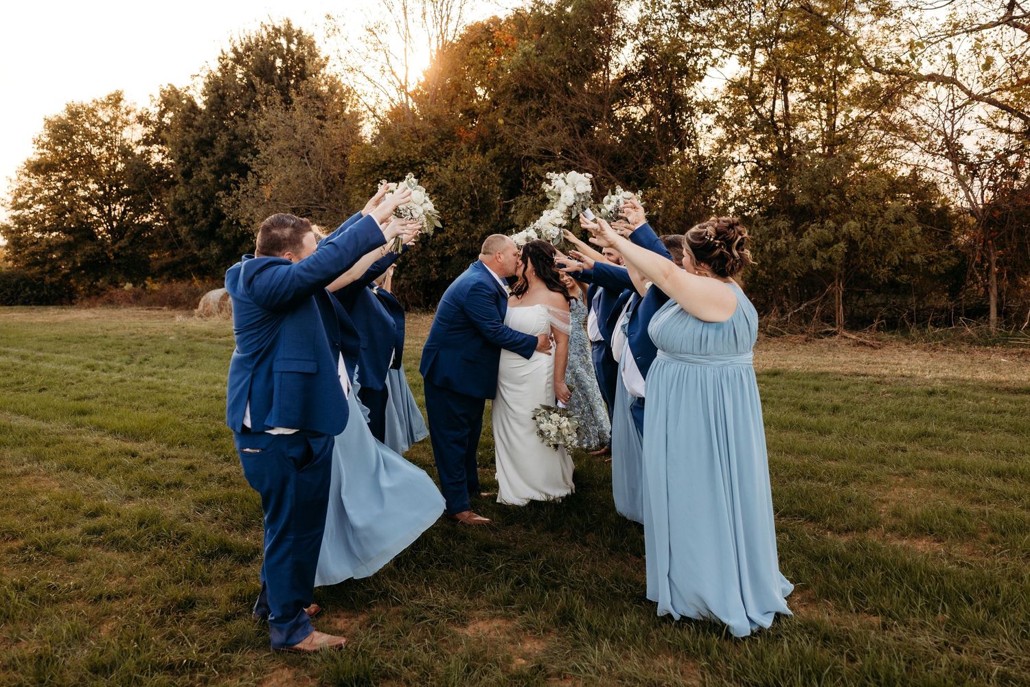 Wedding party celebrates outdoors at golden hour, bridesmaids in blue dresses raising bouquets over bride and groom.