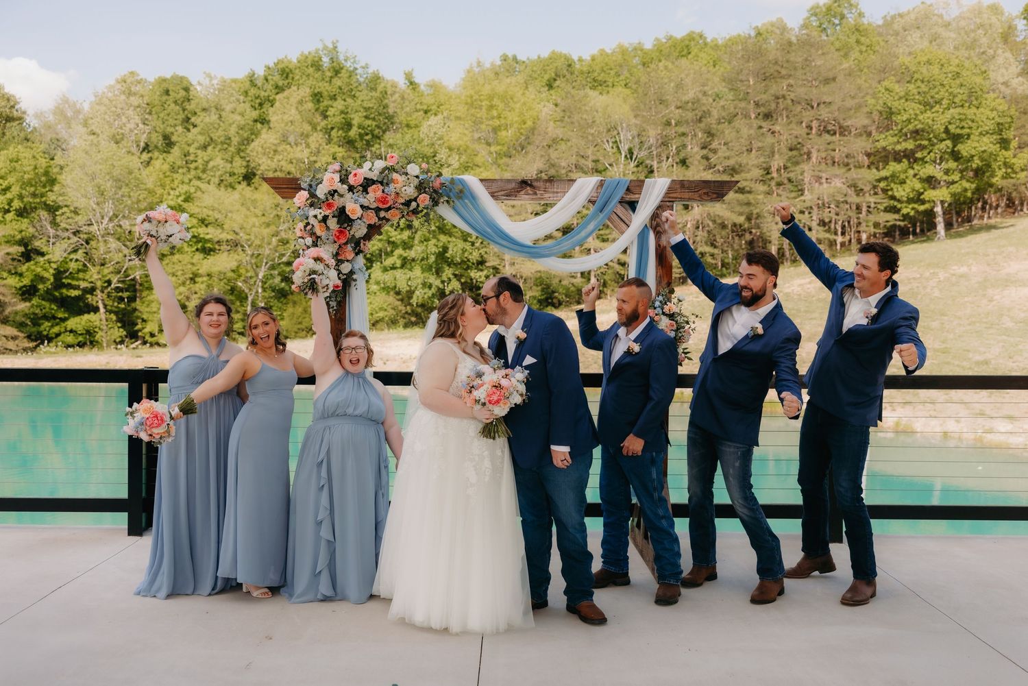 Wedding party celebrates under floral arch with bridesmaids in blue dresses and groomsmen in navy suits.