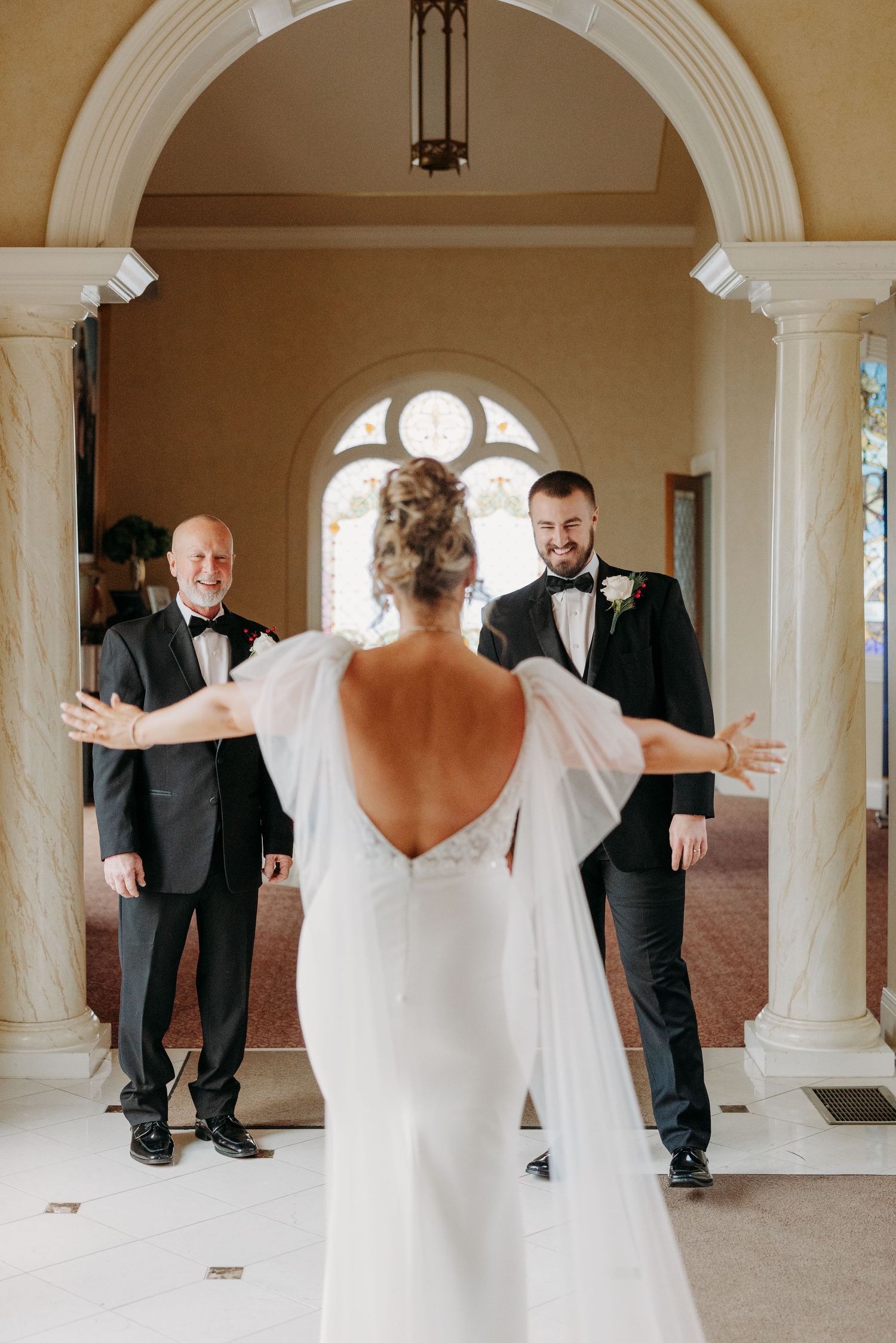 Bride in backless white gown with cape sleeves shares first look moment with groom and father in elegant venue.