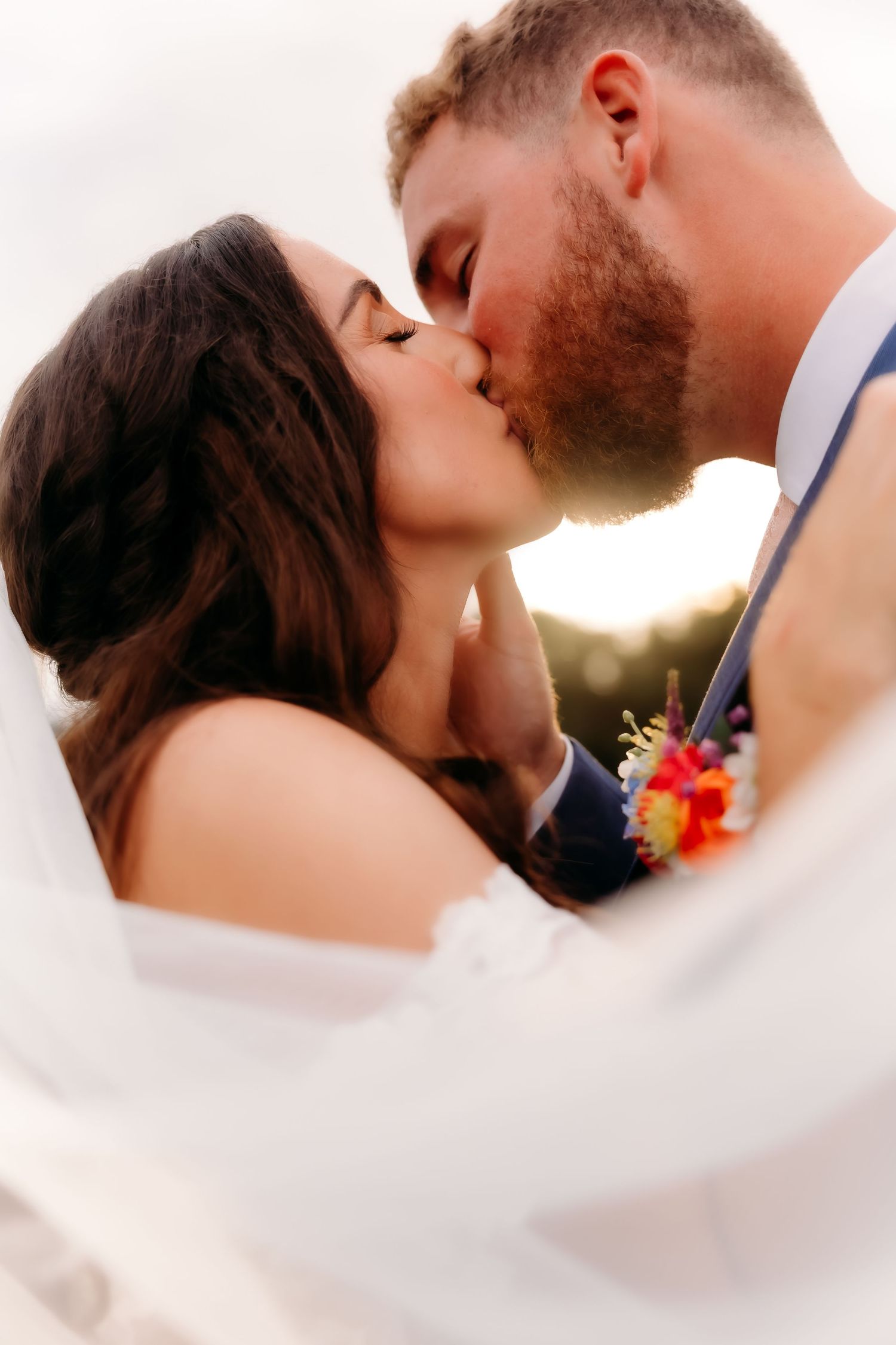 Bride and groom sharing a romantic golden hour kiss, groom wearing a colorful boutonniere, veil flowing softly.