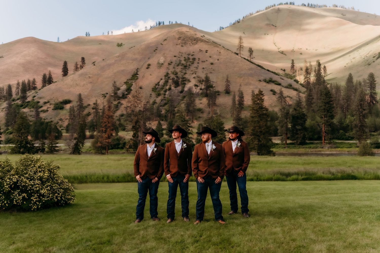 Four groomsmen in matching brown suits and cowboy hats stand together on green lawn with rolling hills backdrop.