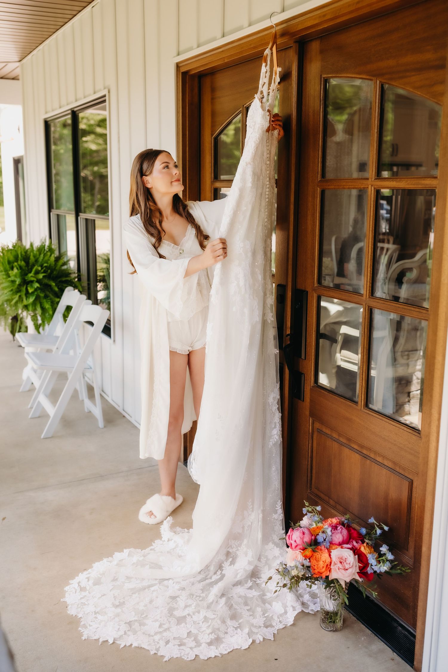 Bride in robe holding up lace wedding dress outside wooden door with colorful floral bouquet nearby.